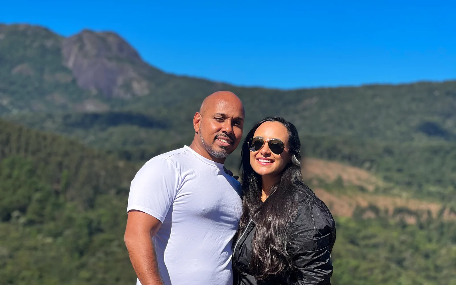 Bárbara e Eduardo sorrindo no mirante, com a serra ao fundo